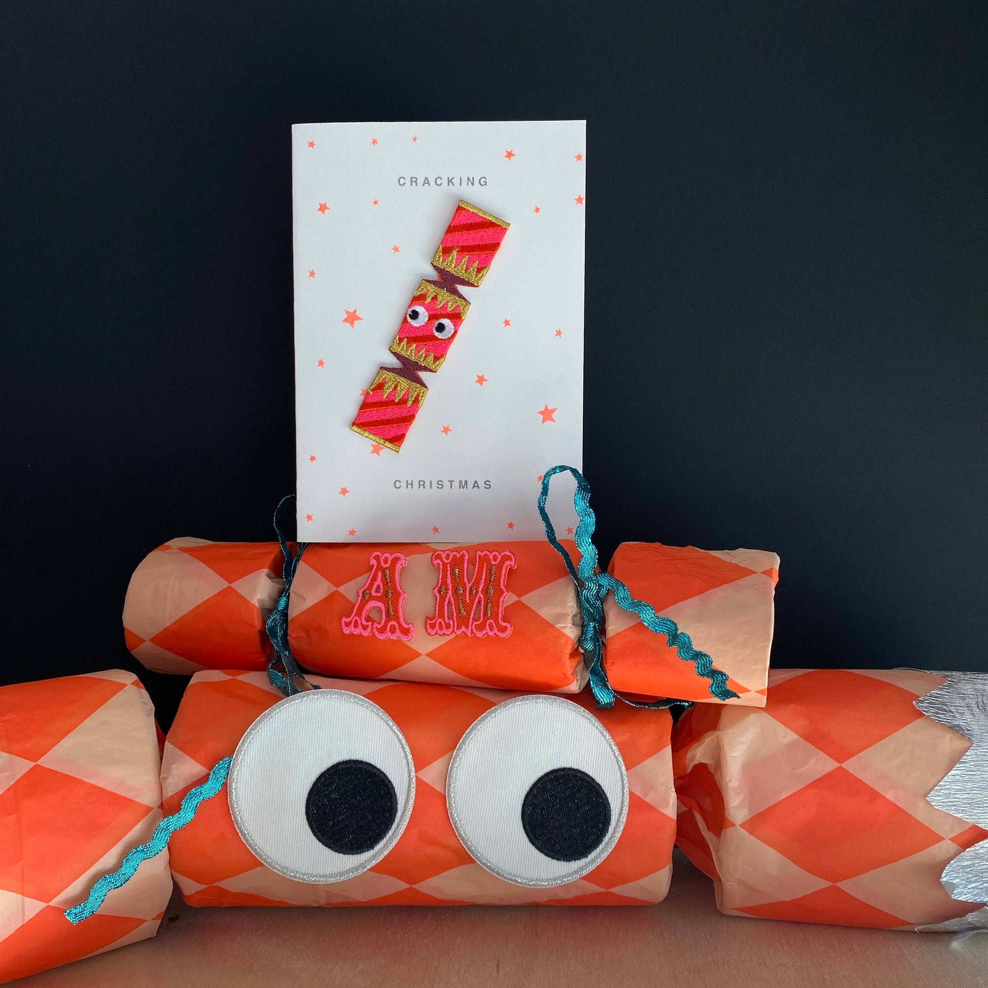  an embroidered Christmas cracker with goggly black eyes.It is pink in colour with purple and golden decoration on the wrapper. The background is white with pink stars. The patch rests on a real life size decorative Christmas cracker with  the letters A and M on it. 