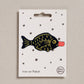 An embroidered iron-on patch shaped like a classic soy sauce fish bottle, featuring a black fish body with gold embroidered details and a bright red bottle cap. Packaged on a cream backing card with black stars and Petra Boase branding.