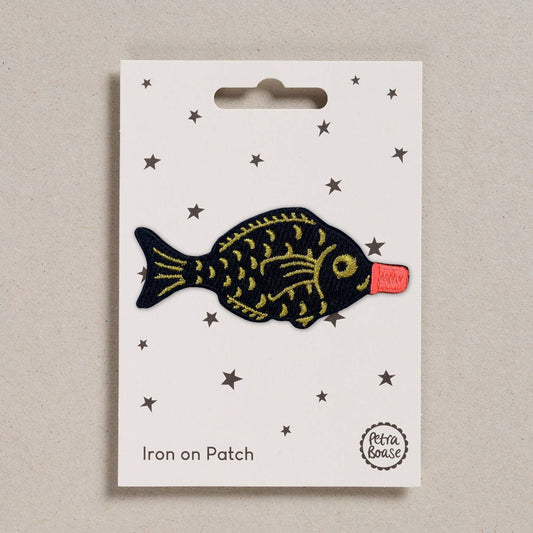 An embroidered iron-on patch shaped like a classic soy sauce fish bottle, featuring a black fish body with gold embroidered details and a bright red bottle cap. Packaged on a cream backing card with black stars and Petra Boase branding.