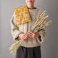 Person wearing a beige sweater holding a bunch of wheat with a yellow patterned wheat bag draped over their shoulder against a gray background.