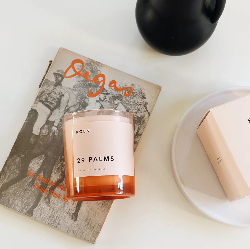 A signature pink candle jar with a pink label with black writing. It sits on a white table on top of a book next to a black vase.