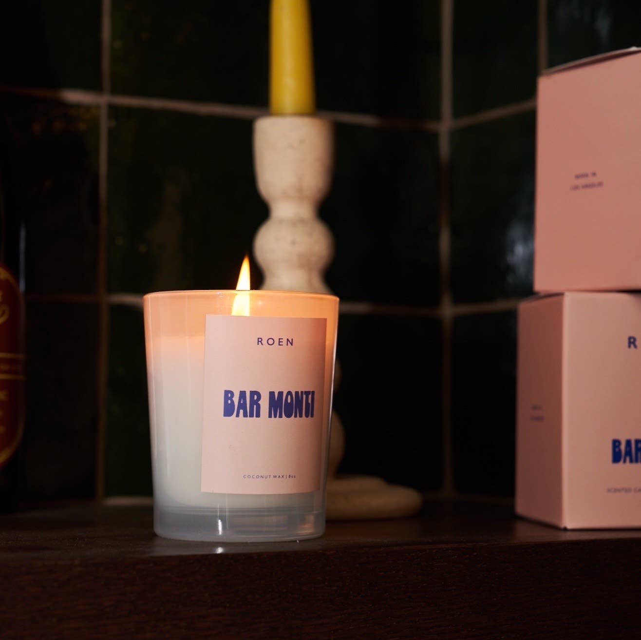 A clear jar with a white candle, with a pink label and blue text stating the band and candle name. It sits against a black tiled wall, next to a couple of the pink boxes that the candles are boxed in.