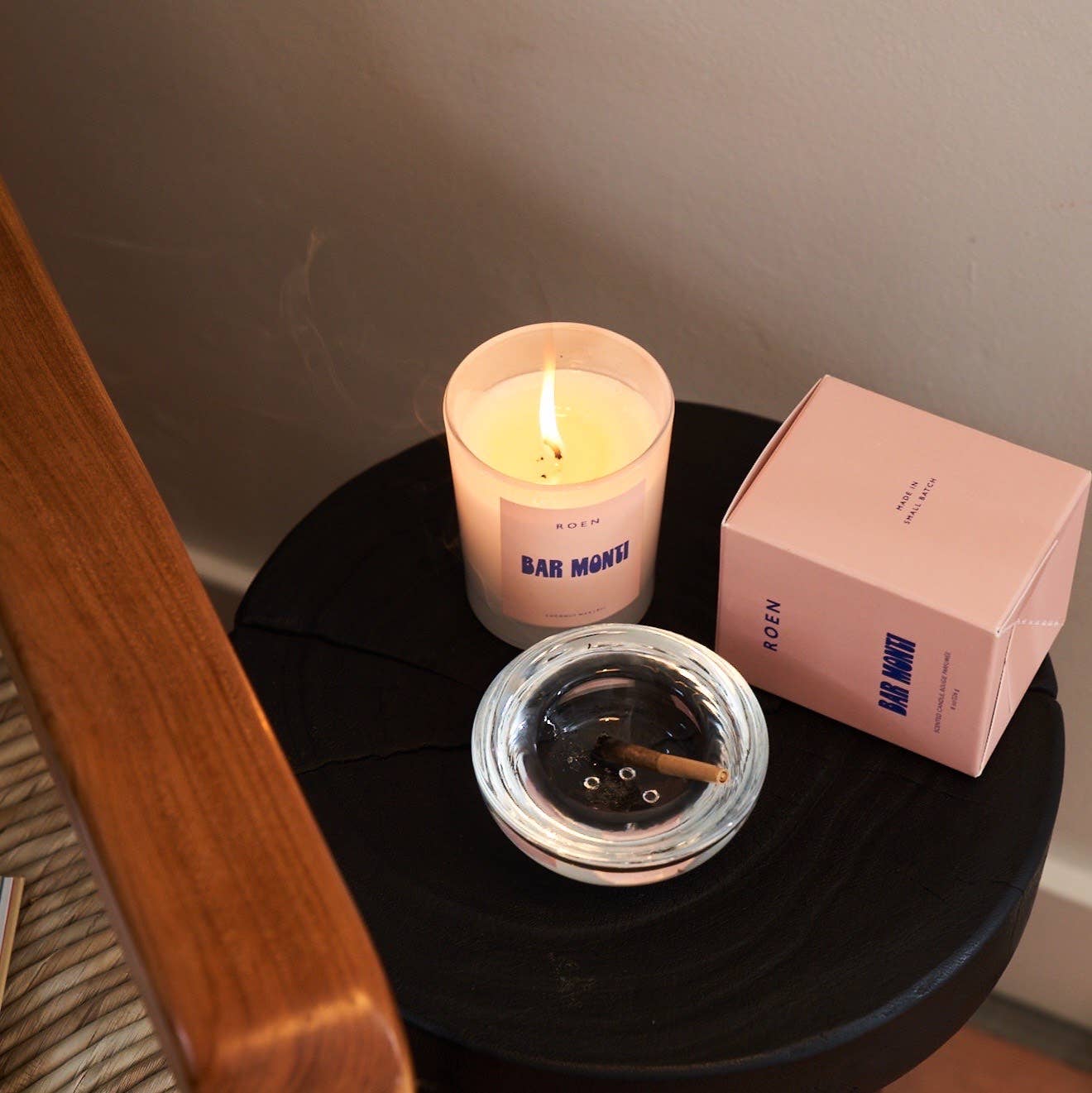 A clear jar with a white candle, with a pink label and blue text stating the band and candle name. It sits on a black side table next to an ashtray, with a pink box that the candles are boxed in.