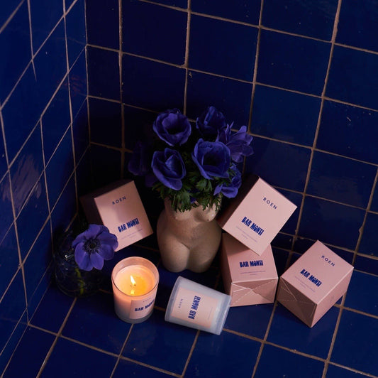 A clear jar with a white candle, with a pink label and blue text stating the band and candle name. It sits against a blue tiled wall, next to a couple of the pink boxes that the candles are boxed in, and a vase containing blue flowers.