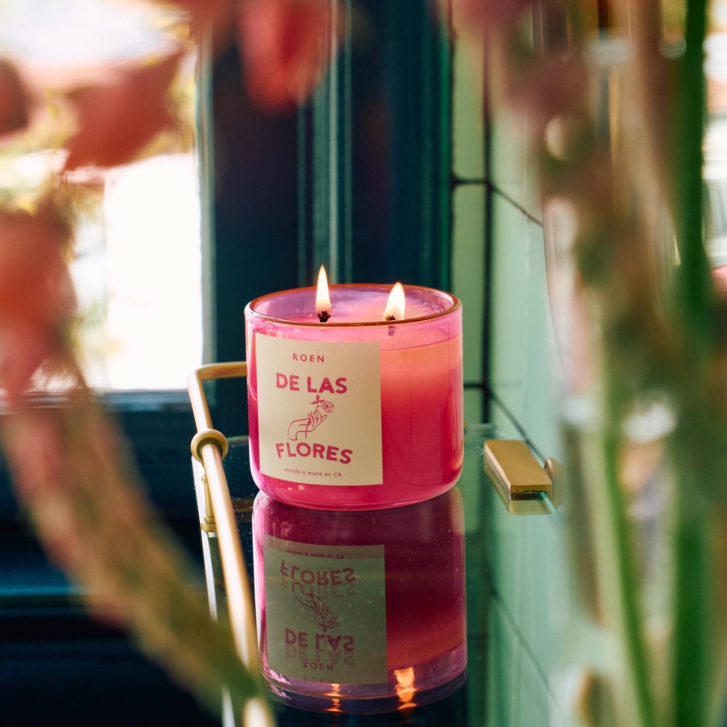 A bright pink jar containing a white candle with two wicks.  white label with bright pink writing. It sits on a mirrored surface with some blooms in the foreground.