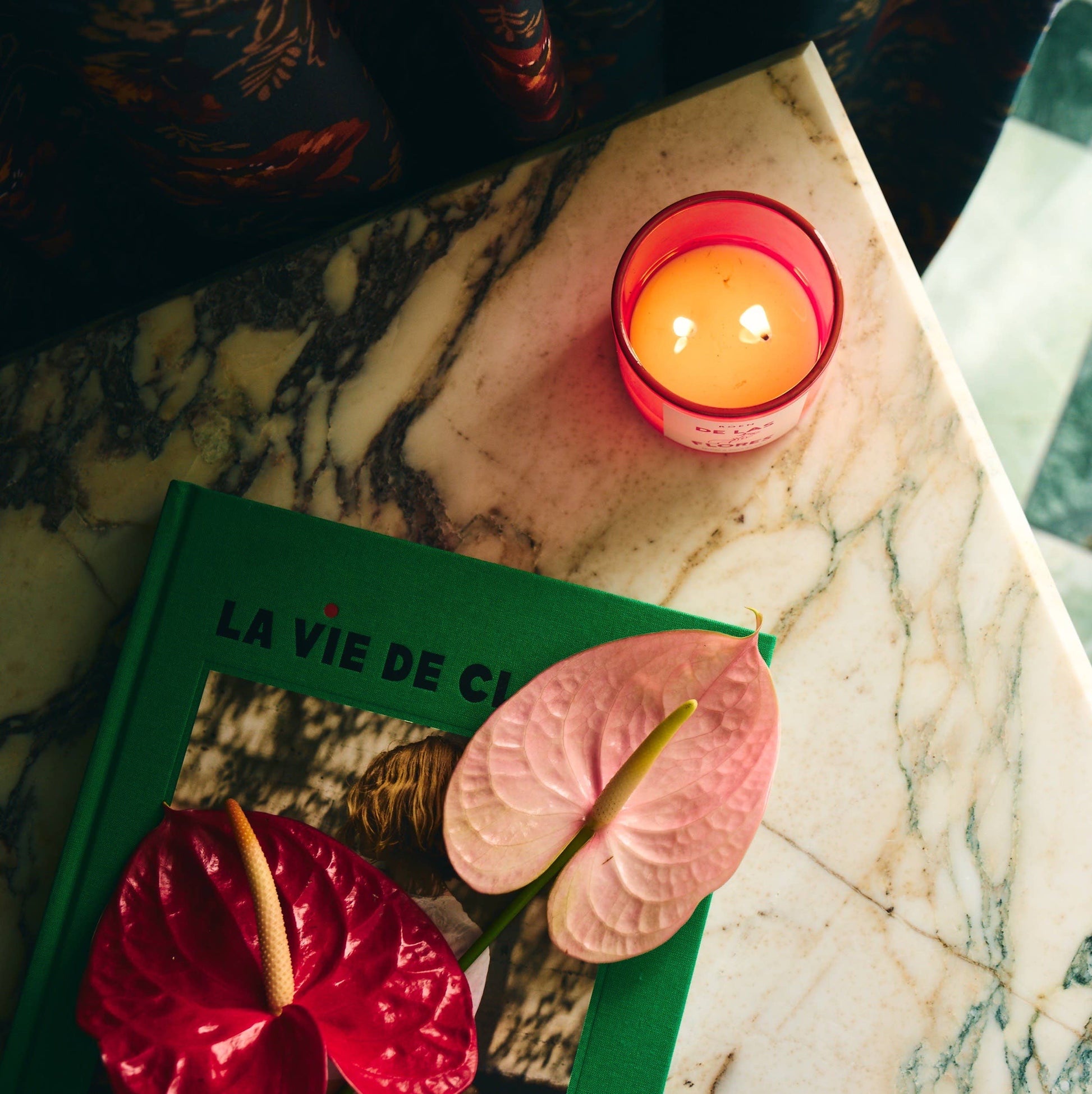 On a marble table sits a bright pink candle which is lit with two wicks. A green book sits next to it with two orchid flowers lying on it.