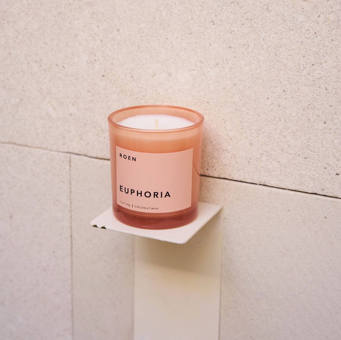 A white candle in a pink glass jar. Te pale pink label has black writing with the brand and candle fragrance name on it.