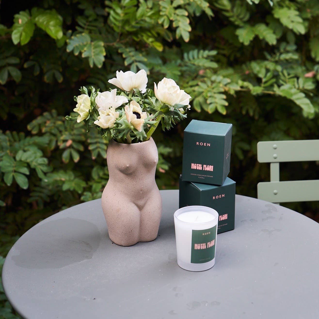 A white jar contains a white candle with a green label with pink writing. Towo green boxes with pink writing sit behind it on a garden table next to a vase containing white flowers 