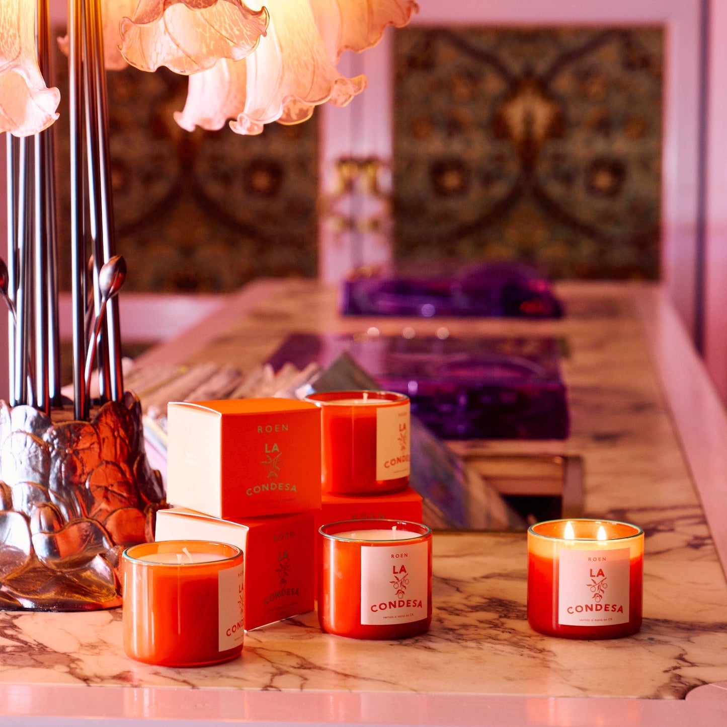 A bright orange glass jar with a contrast turquoise rim. The candle is white with 2 wicks. The label is white with orange writing. There are three and thy sit in an exotic looking setting with a marble tabletop and soft lighting. There are orange boxes 