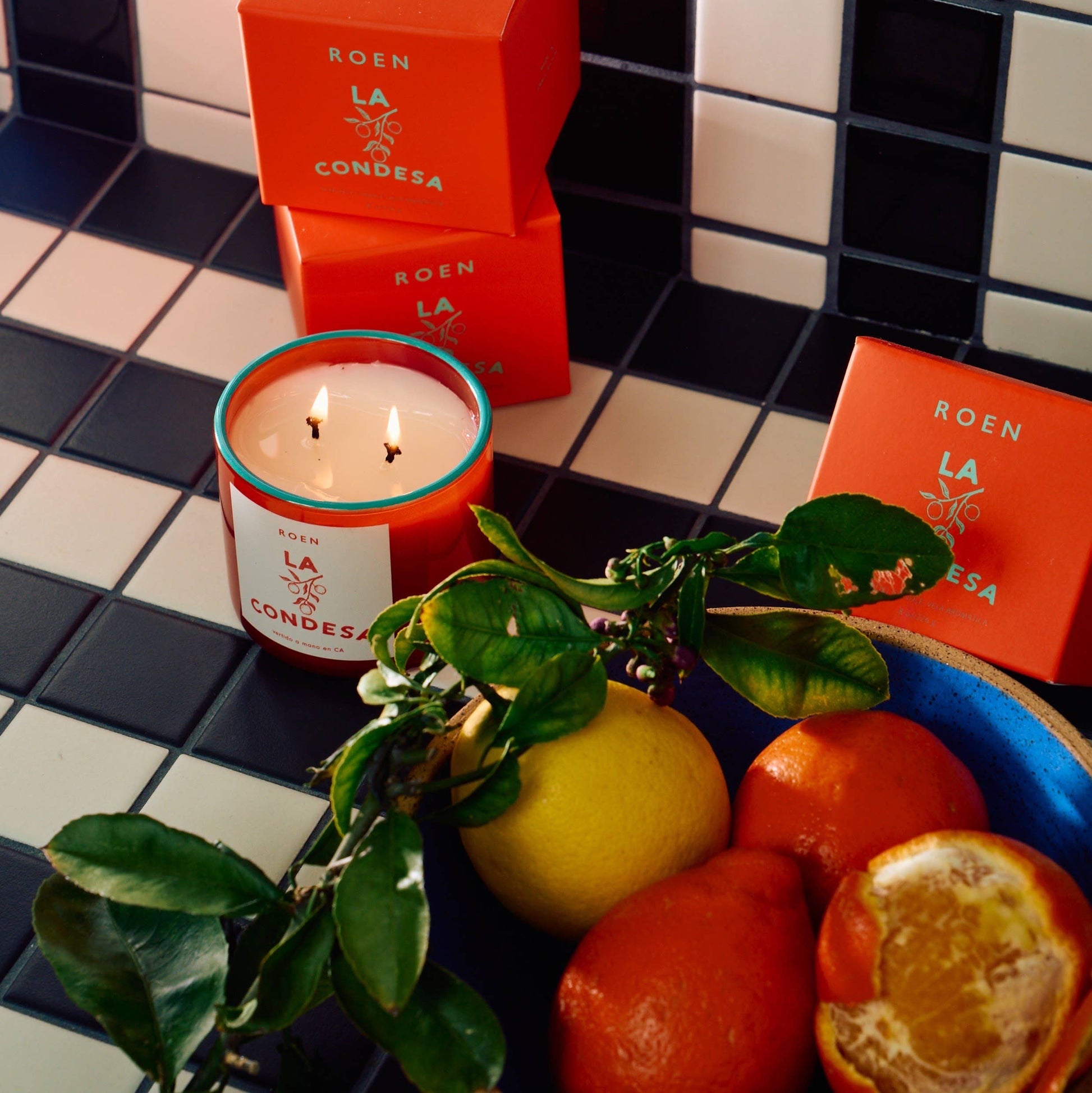 A bright orange glass jar with a contrast turquoise rim. The candle is white with 2 wicks. The label is white with orange writing. The candle is lit and sits on a black and white tiled surface next to a fruit bowl containing citrus fruit. The orange boxes of the candles are also shown.