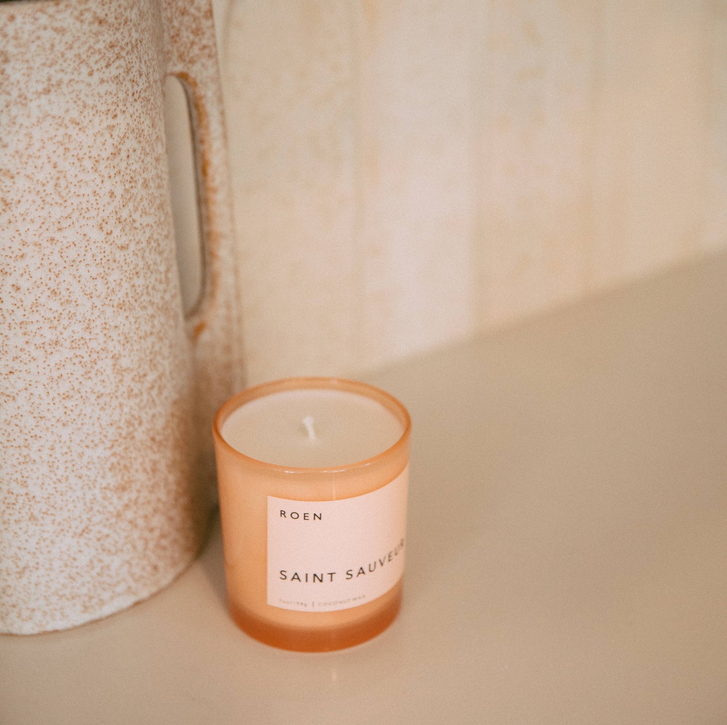 A pink glass jar with a white label with black text stating the brand name and candle fragrance name. It sits on a cream marble worktop against a beige speckled jug.