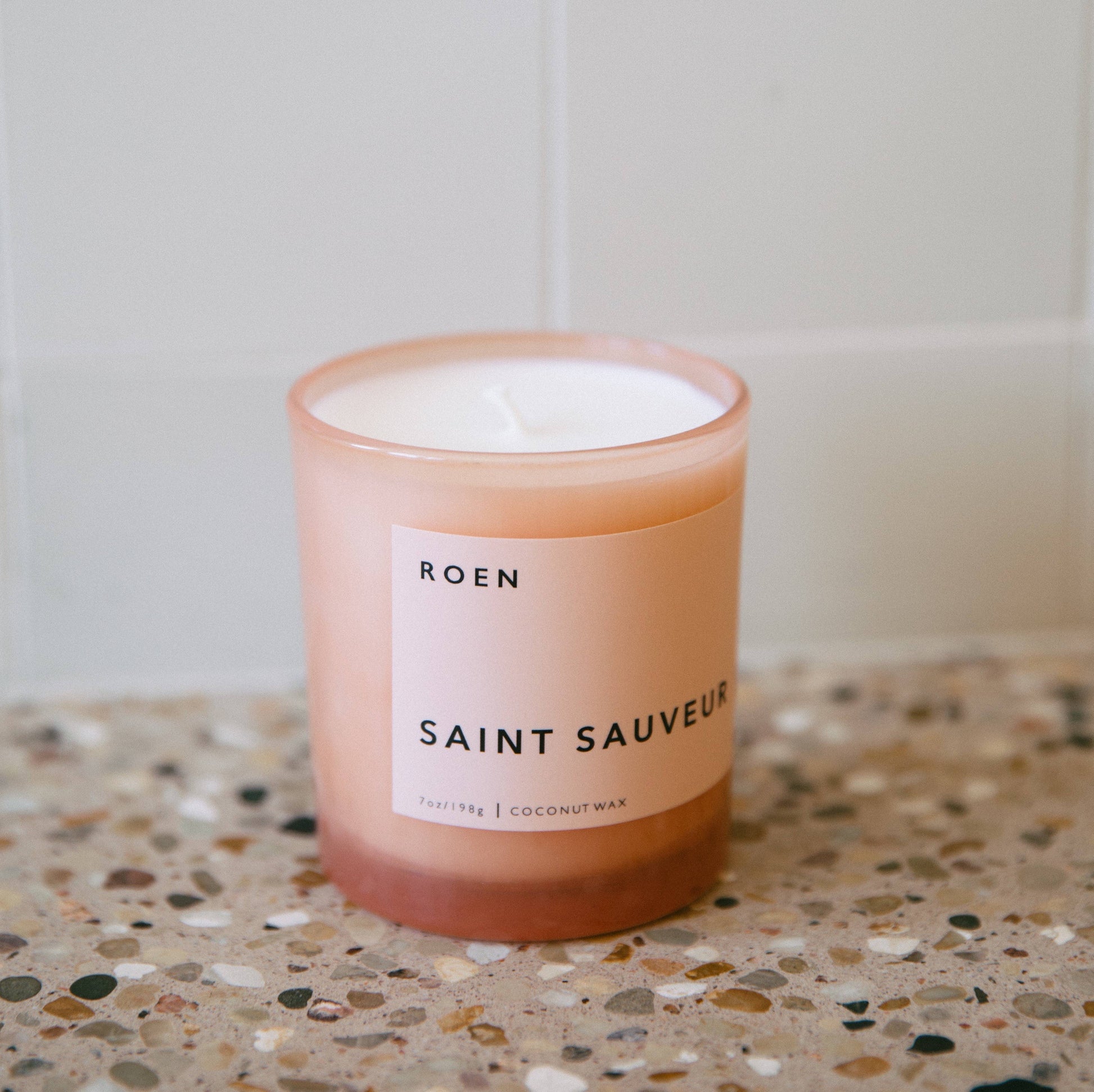 A pink glass jar with a white label with black text stating the brand name and candle fragrance name. It sits on a terrazzo worktop