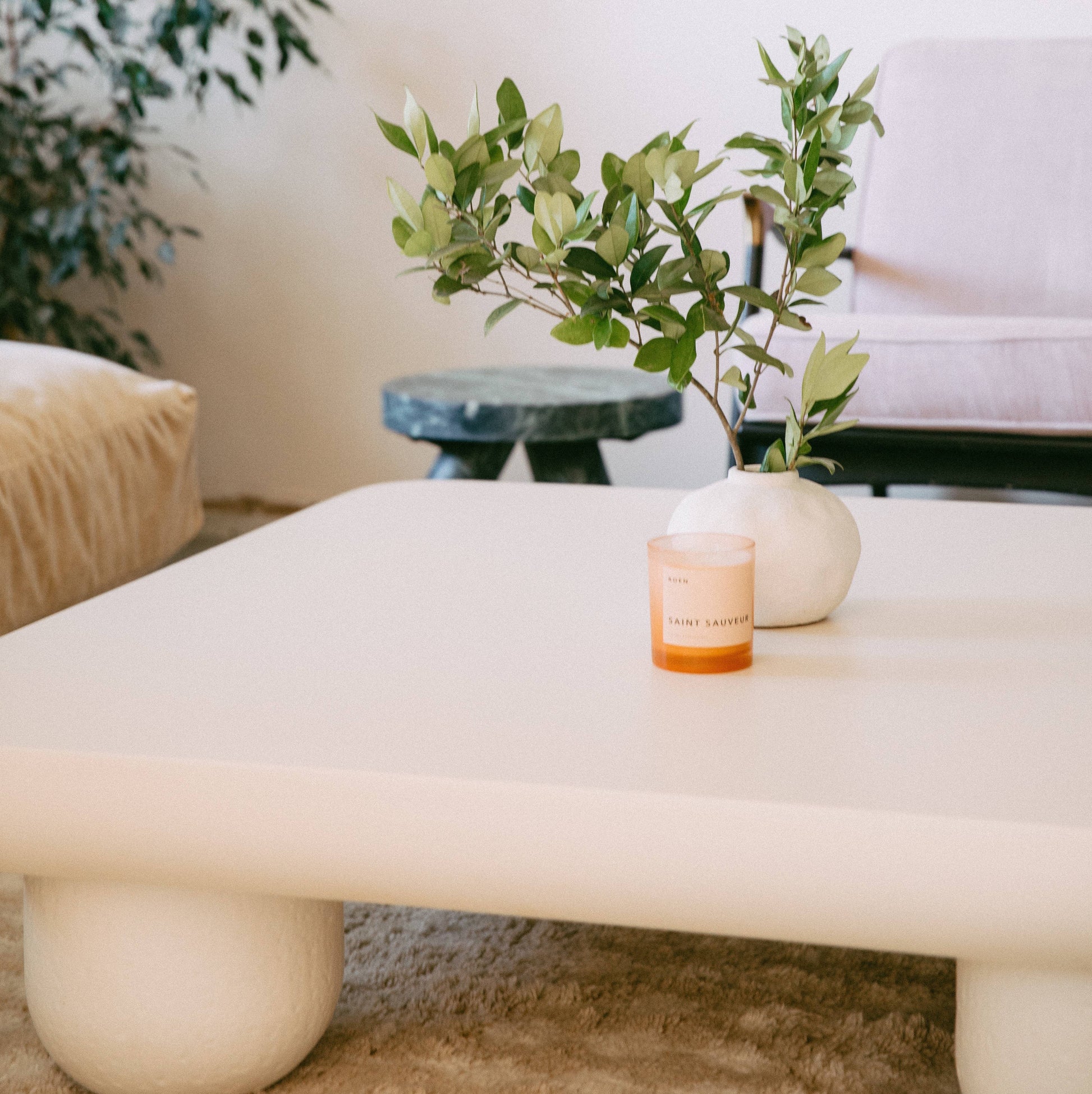 A pink glass jar with a white label with black text stating the brand name and candle fragrance name. It sits on a marble coffee table in front of a cream vase with green sprigs of foliage.