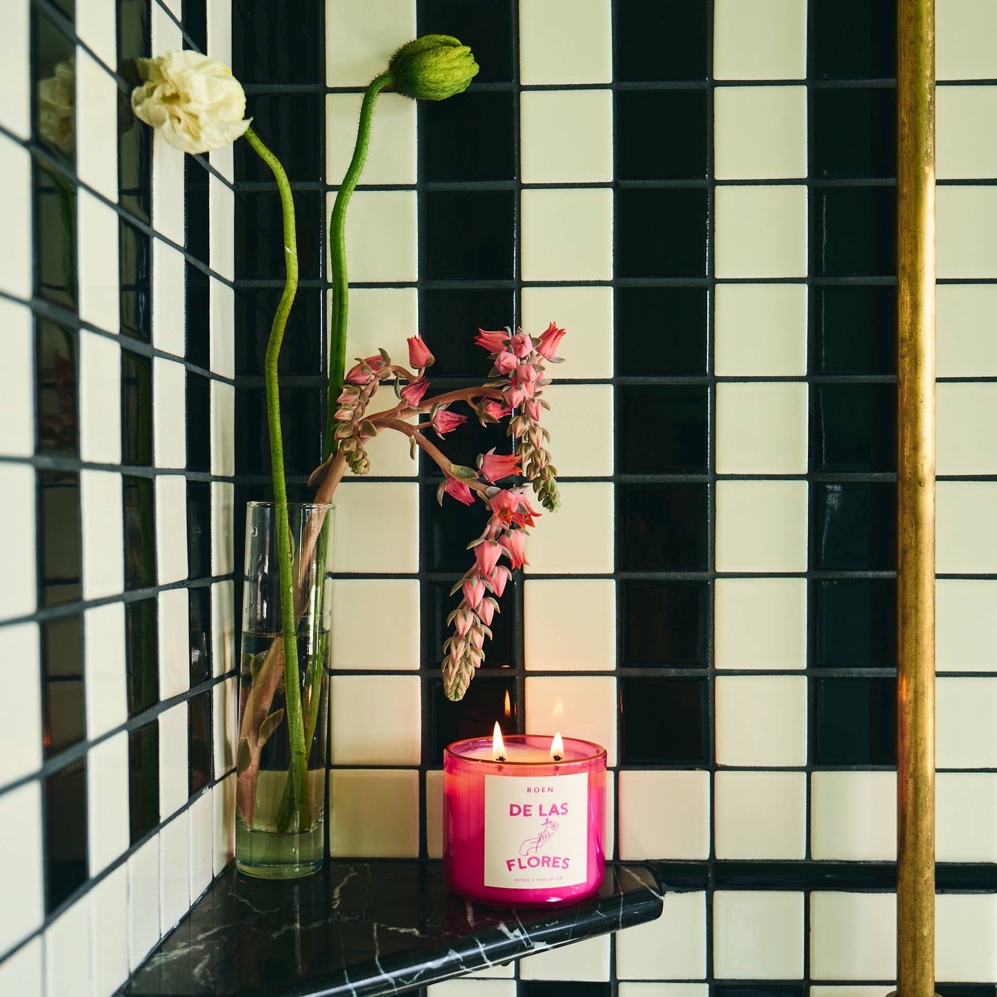 A bright pink jar containing a white candle with two wicks.  white label with bright pink writing. It sits in a black and white tiled room  with some blooms in a vase in the background.