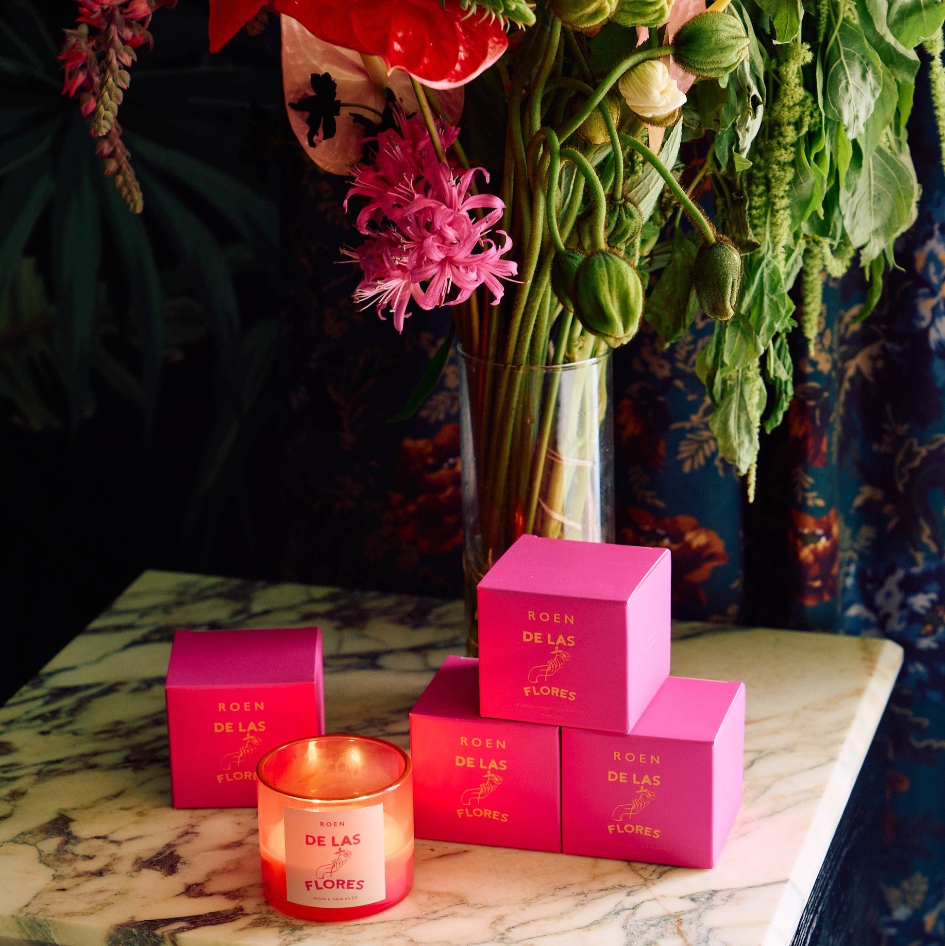 On a marble table sits for bright pink boxes containing candles. One sits in font of them - it has a bright pink jar with a white label with pink writing. It is lit and burns with two wicks. In the background is a large clear vase contains some flowers.