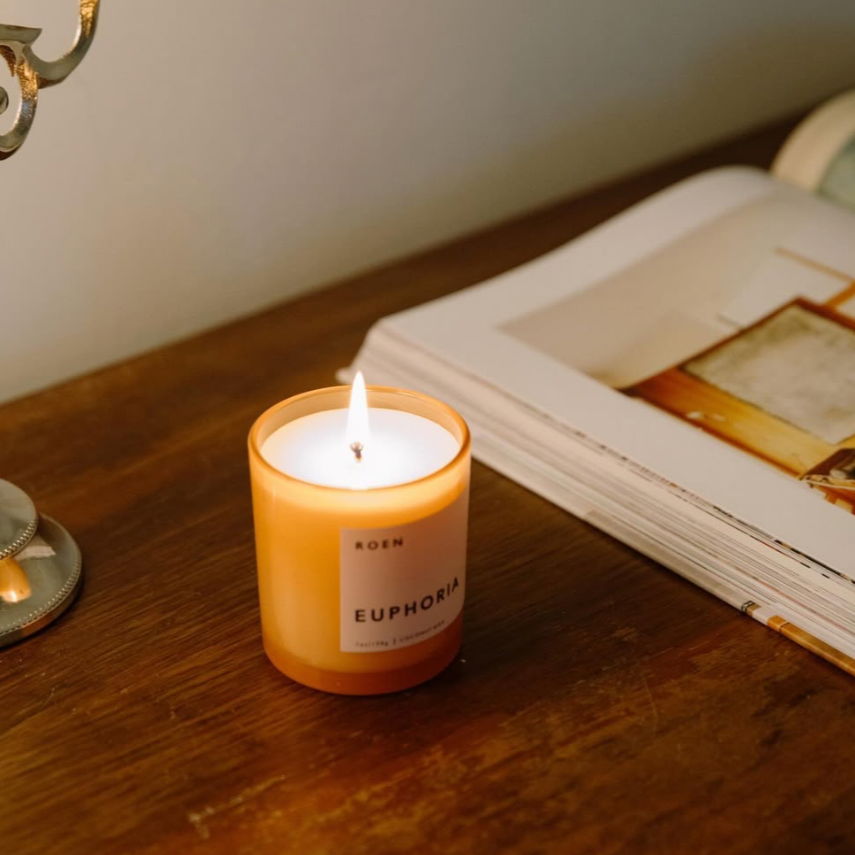 A signature pink candle jar with a pink label with black writing. It is lit with a gentle flame and sits next to an open book