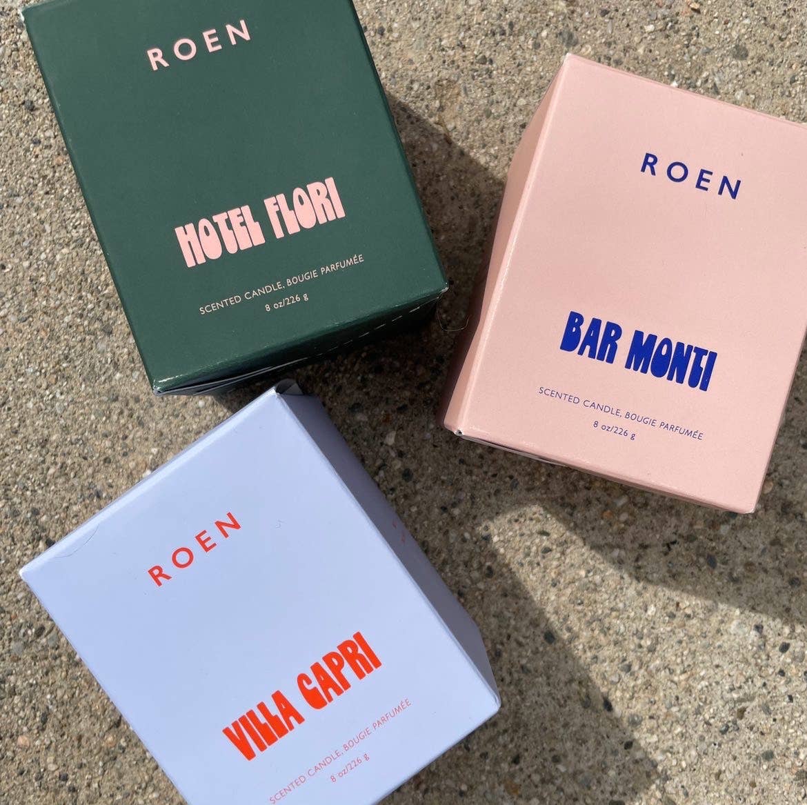 Against a concrete surface, the three boxes of the Roen candle collection sit. One is green with pink writing, one is pink with blue writing, and one is blue with red writing. 