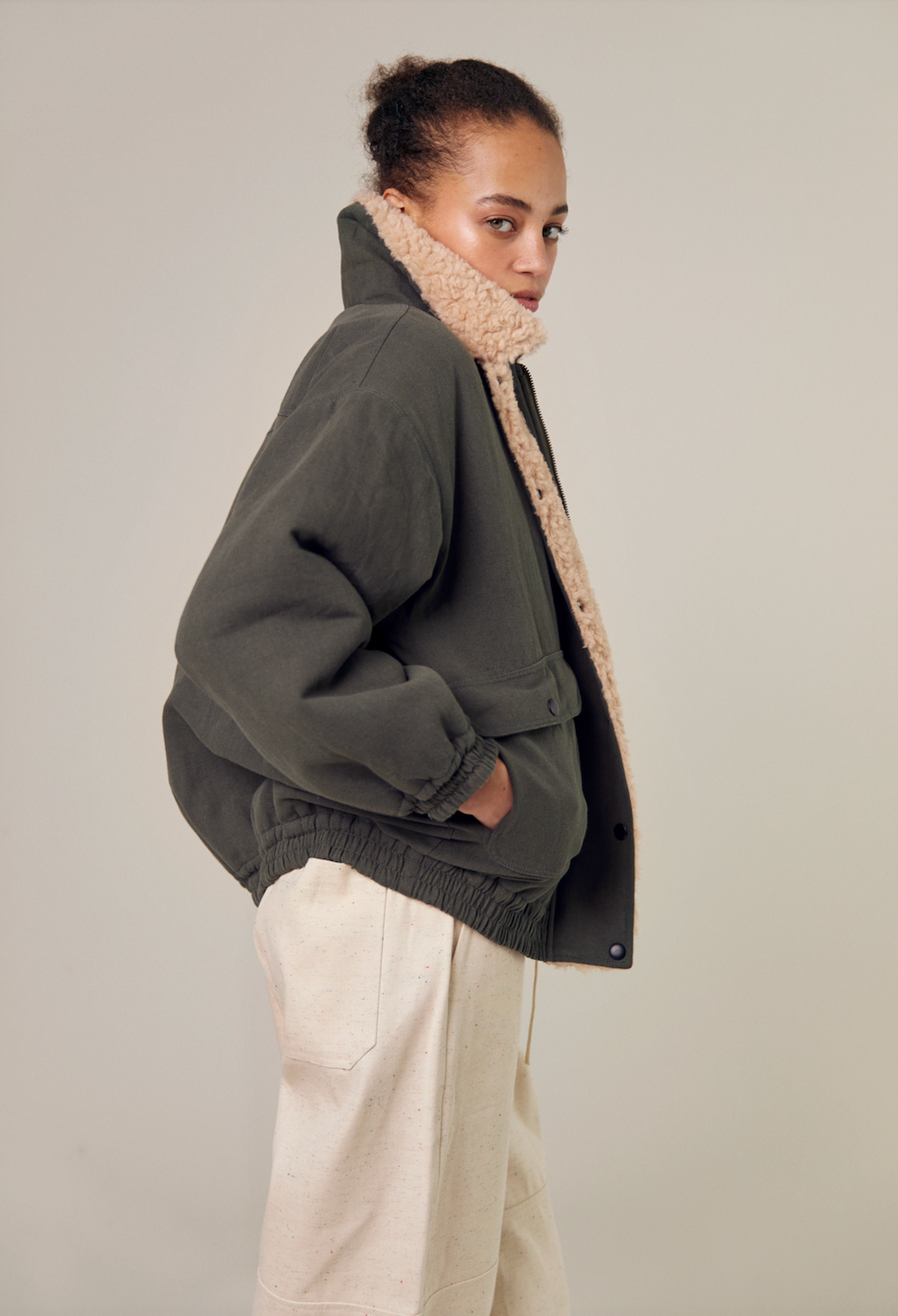 A side view of the Sideline Wilma Jacket, featuring the olive cotton twill exterior with the collar styled up for added elegance. The model showcases the sleek silhouette, highlighting the zip front closure and the rounded collar. This angle emphasizes the jacket's stylish design and practicality, perfect for winter wear.