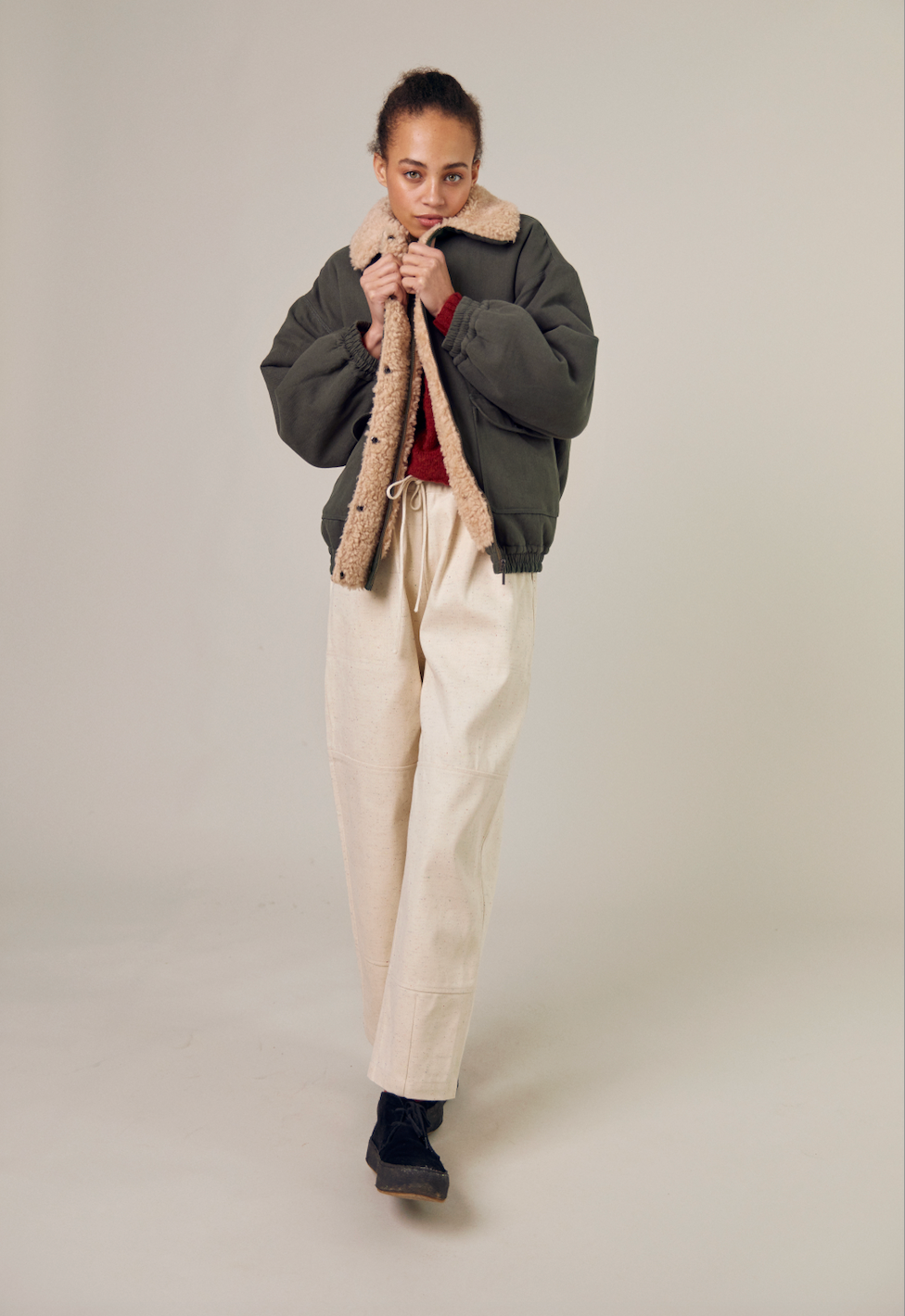 A full view of the Sideline Wilma Jacket, worn by a model holding the jacket closed near her face. She pairs the olive cotton twill jacket with stylish white jeans, creating a chic winter look. The jacket features a zip front closure, snap-button placket, and rounded collar, showcasing its elegant design and versatile appeal.