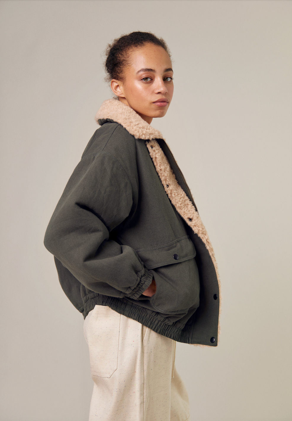 A side view of the Sideline Wilma Jacket, showcasing the olive cotton twill exterior. The jacket features a zip front closure and rounded collar, with the model displaying its sleek silhouette. The design highlights the jacket's stylish lines and practical elements, making it a versatile choice for winter wear.