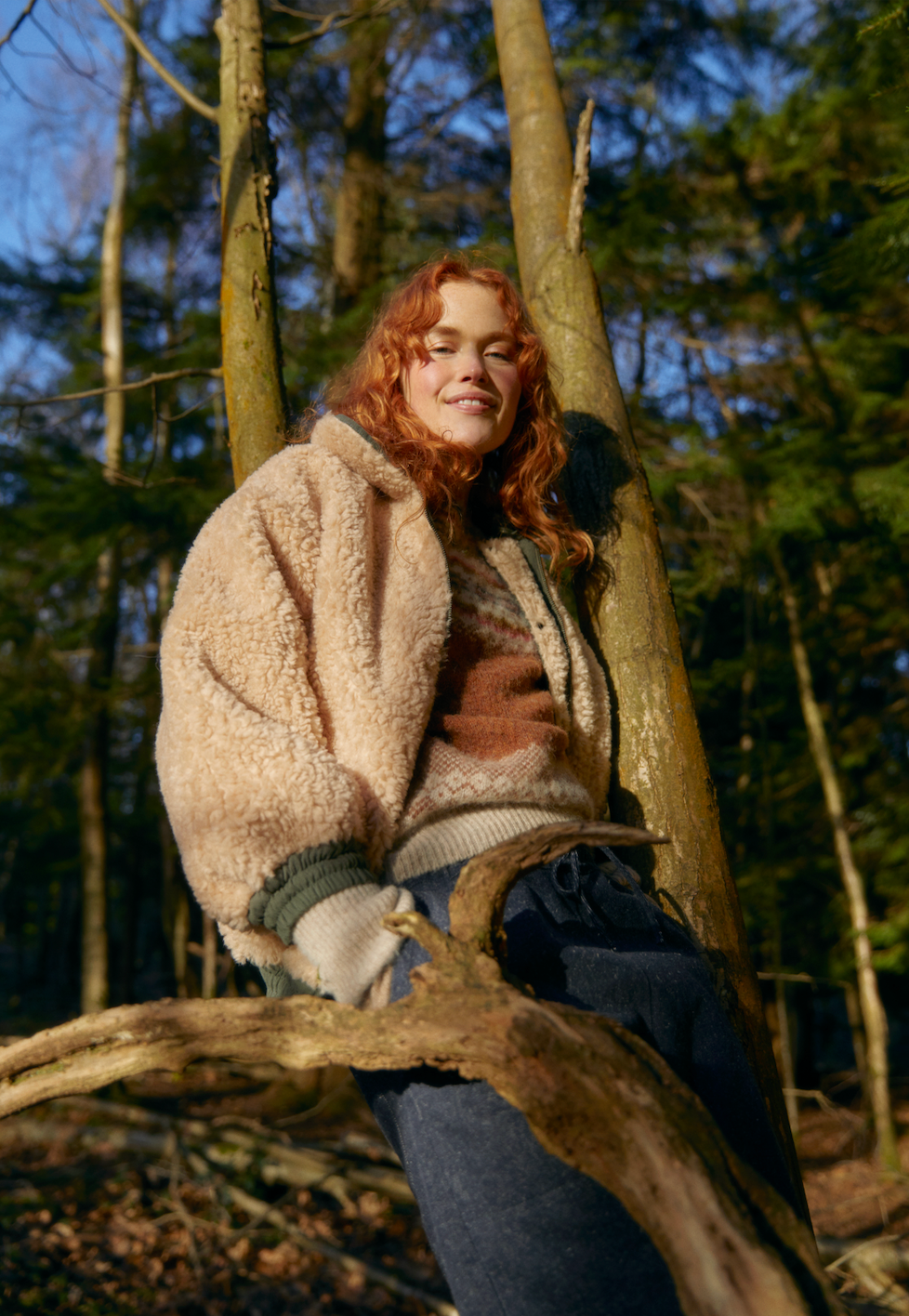 A campaign shot of the Sideline Wilma Jacket, featuring the olive cotton twill side, as the model sits against a tree in a beam of sunlight. She smiles warmly while wearing a cozy knit sweater and rolled-up jeans, embodying the spirit of winter comfort and style amidst a natural setting.