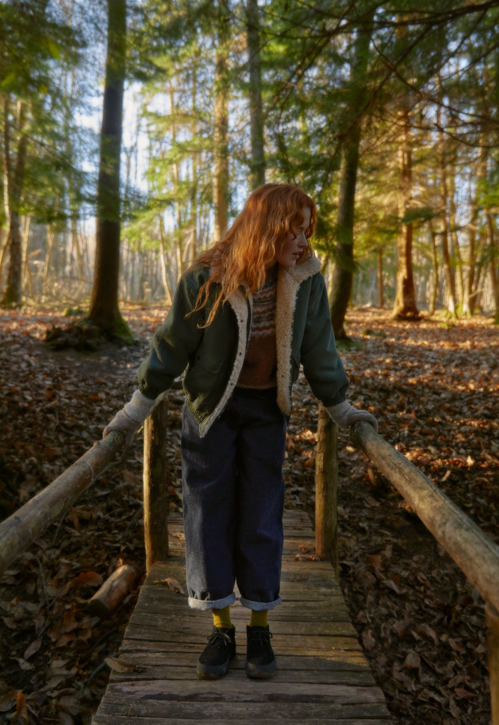 A campaign shot of the Sideline Wilma Jacket featuring the olive cotton twill side, worn by a long-haired curly model in the woods on a wooden pathway. She pairs the jacket with a cozy knit sweater, rolled-up jeans, and stylish ankle boots, creating a relaxed yet chic look that embodies winter warmth and outdoor adventure.