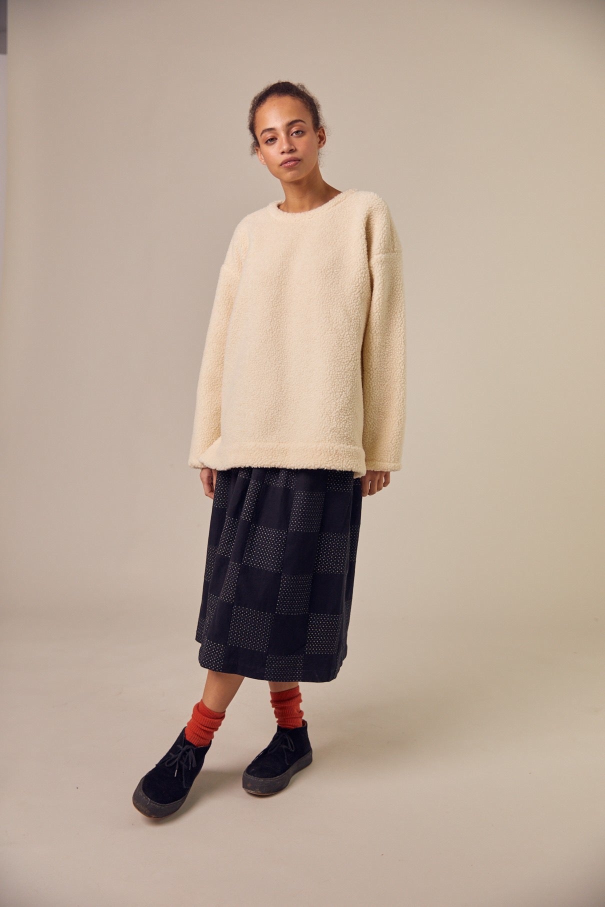 The Sideline Rosa Top, styled on a model who pairs it with a long checked skirt, red ankle socks, and ankle boots. The oversized shearling sweatshirt adds warmth and comfort, while the outfit showcases a trendy mix of textures and patterns, perfect for a stylish winter look.