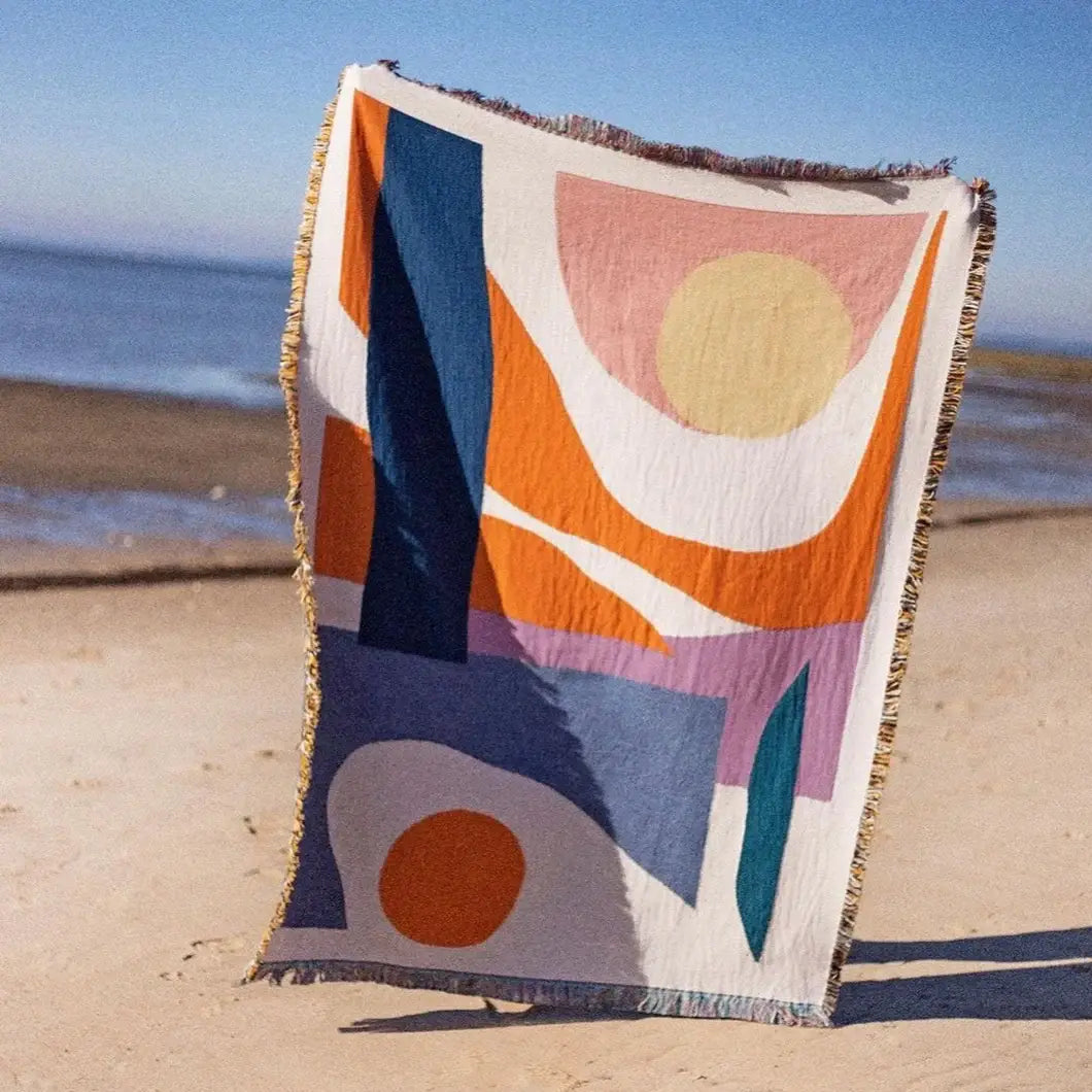 The Essien Throw held up on a sandy beach, its woven fabric catching the light, making it a perfect lightweight throw for picnics and outdoor lounging.