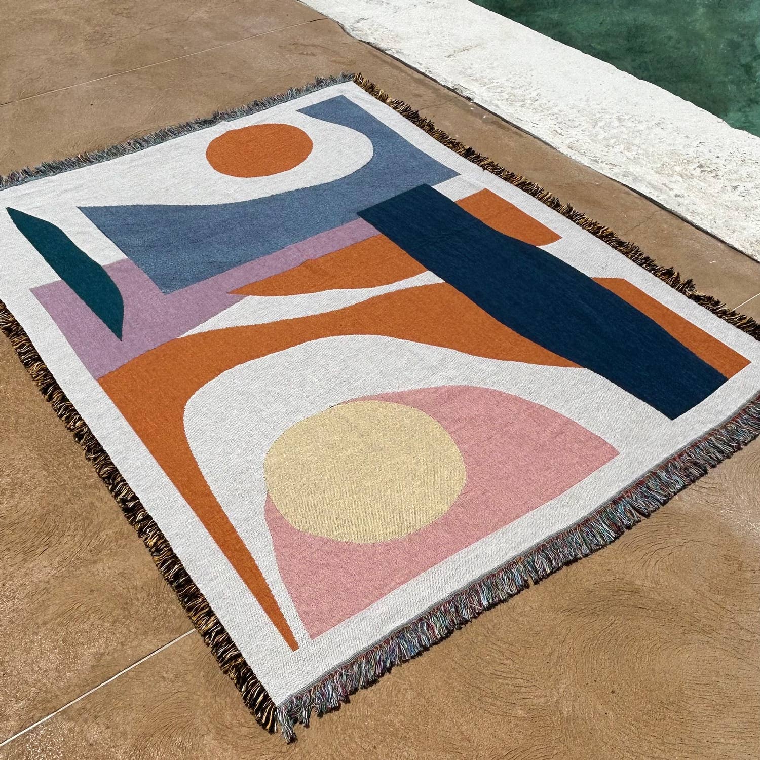 The Essien Throw laid out by a poolside, showing its lightweight, woven texture and vibrant colour palette. Designed for indoor and outdoor use.