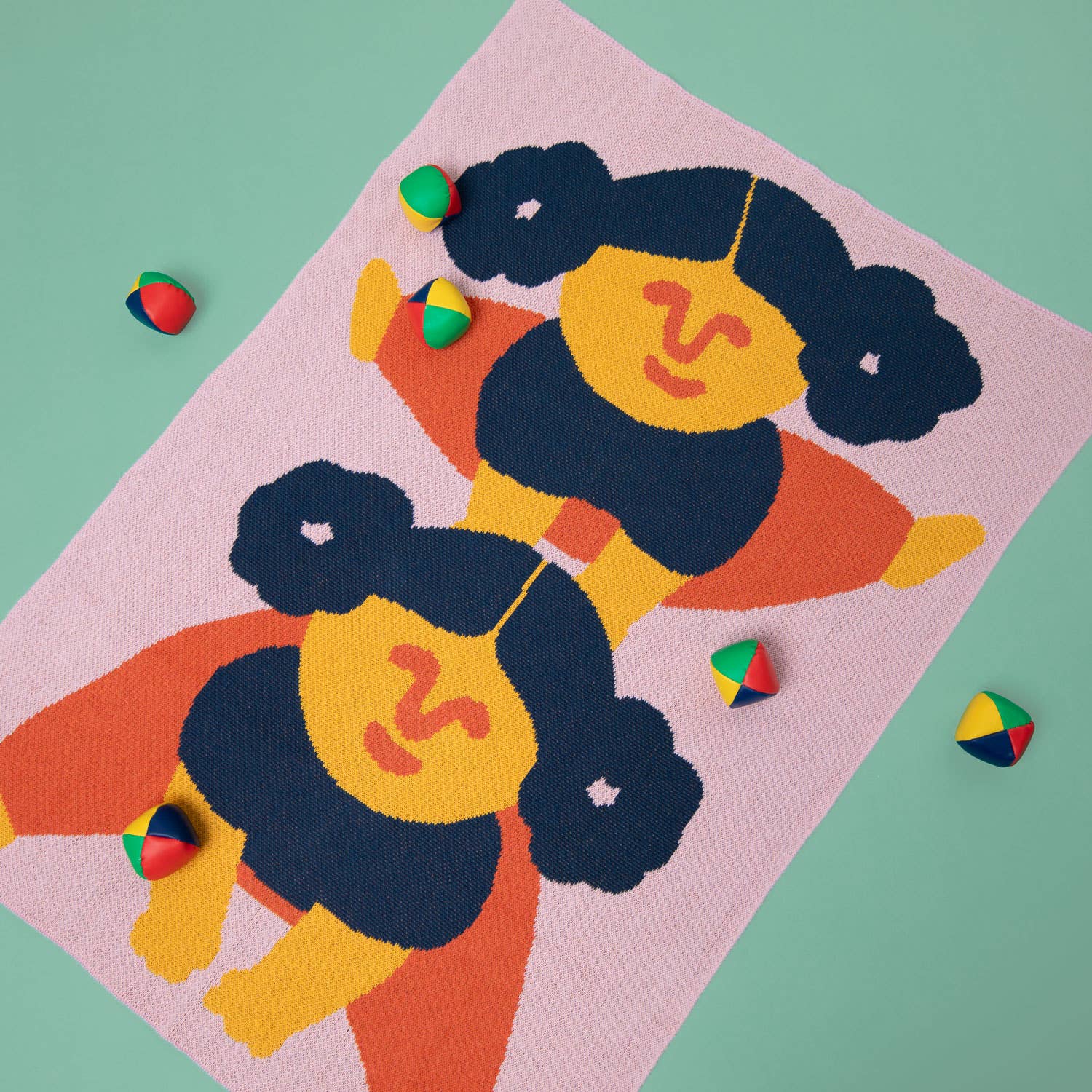 The Leapfrog Mini Blanket, laid flat on a pastel green background, featuring a playful, abstract design in pink, navy, orange, and yellow. Small colourful beanbags surround the blanket.
