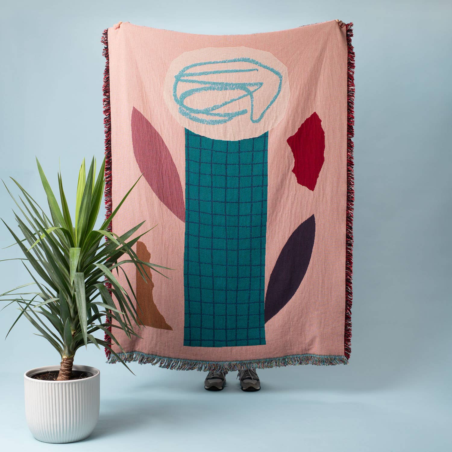 A person holds up the Slowdown Studio Shelley Throw against a light blue background, showcasing its bold abstract design inspired by organic shapes and floral elements. The throw features a mix of blush pink, teal, deep red, and earthy tones, with a striking grid pattern running vertically. A potted plant adds a natural touch to the composition.