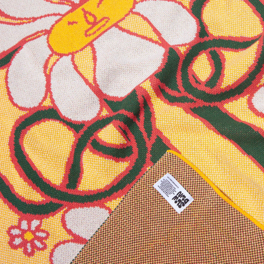 Close-up detail of the knitted daisy blanket, highlighting the intricate weave, bold colours, and texture, with a visible woven label in the corner.