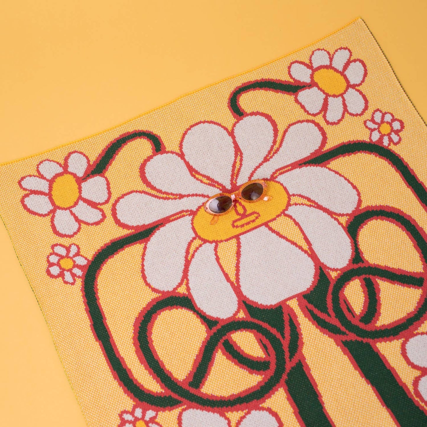 A playful display of the daisy blanket with sunglasses placed on the centre daisy's face, against a bright yellow background.