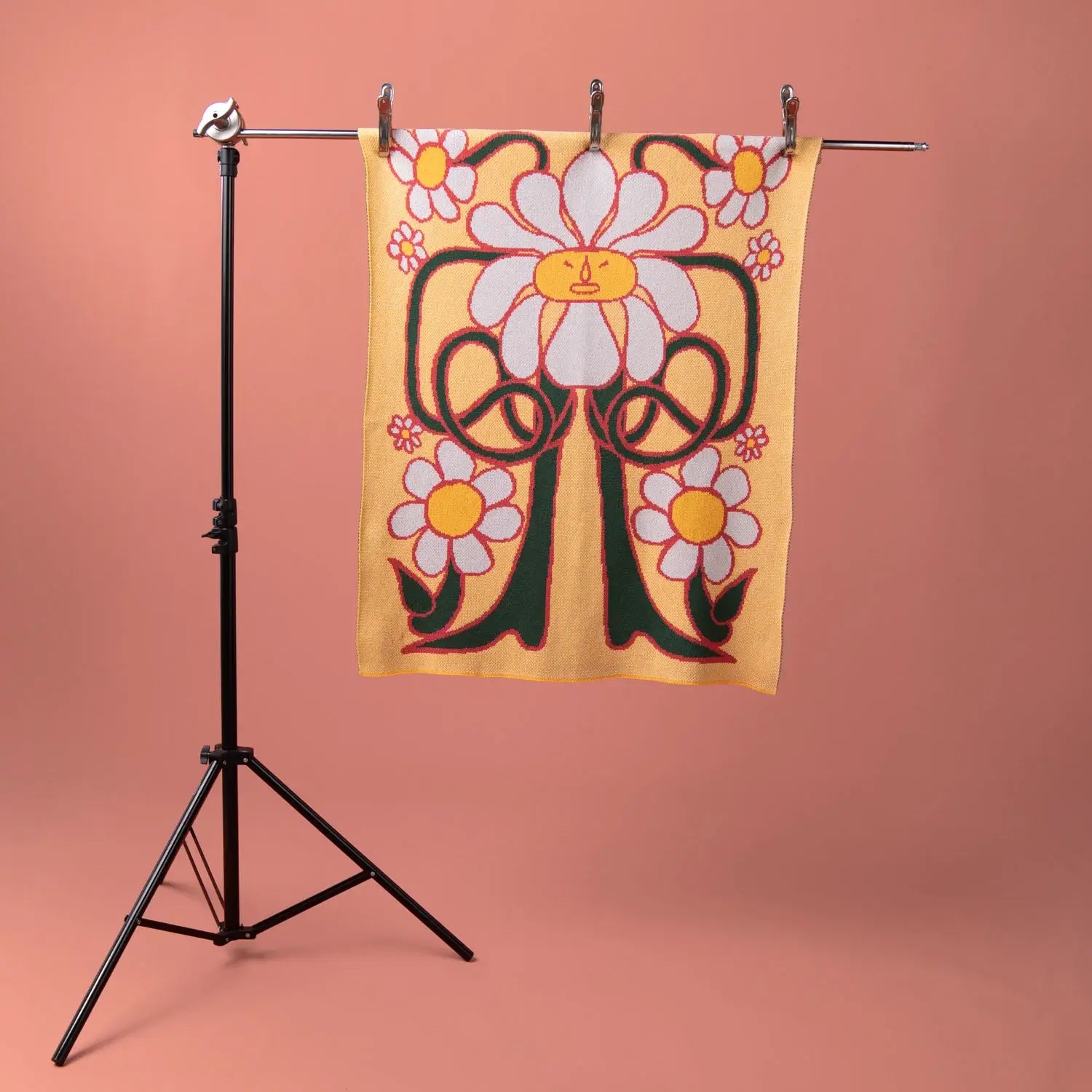 Knitted daisy-patterned blanket in warm yellow tones, hanging on a minimalist black stand with a peach-coloured background, showcasing its full design.