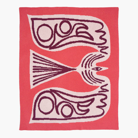 The Slowdown Studio Squiggle Bird Mini Blanket, designed by Marcello Velho, featuring a bold abstract bird in deep berry and blush on a coral background. A soft, woven blanket made from recycled cotton.