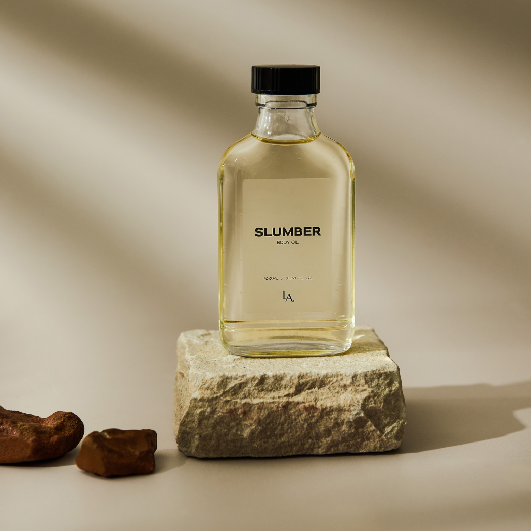 A glass bottle of essential oil blend featuring Rosemary, Frankincense, and Clary Sage, with a black lid and black writing, showcasing translucent oil. The bottle is elegantly positioned on a piece of stone against a neutral background, highlighting its calming properties for a tranquil night's sleep.