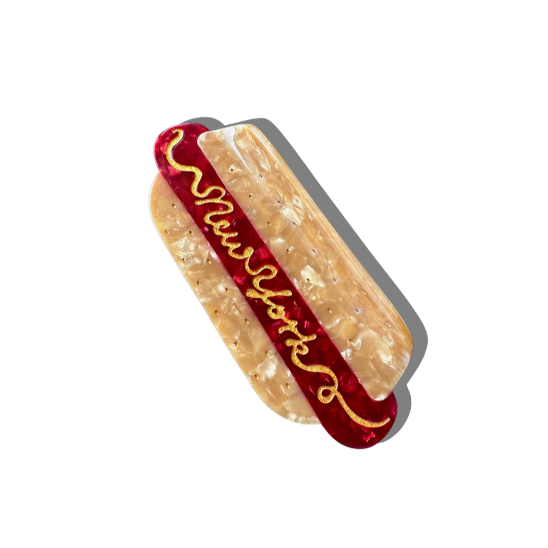 A hot dog-shaped hair claw featuring a marbled beige bun and deep red sausage, finished with gold detailing. The word 'New York' is written in mustard-like script across the top, adding a playful touch. The clip casts a soft shadow against a white background.