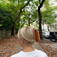 A woman with blonde hair styled in a French twist wears the hot dog-shaped hair claw, securing her updo with a fun and quirky accessory. The marbled beige and red design stands out against her white blouse. She is walking along a tree-lined pathway covered in autumn leaves, with city streets and parked cars in the background.