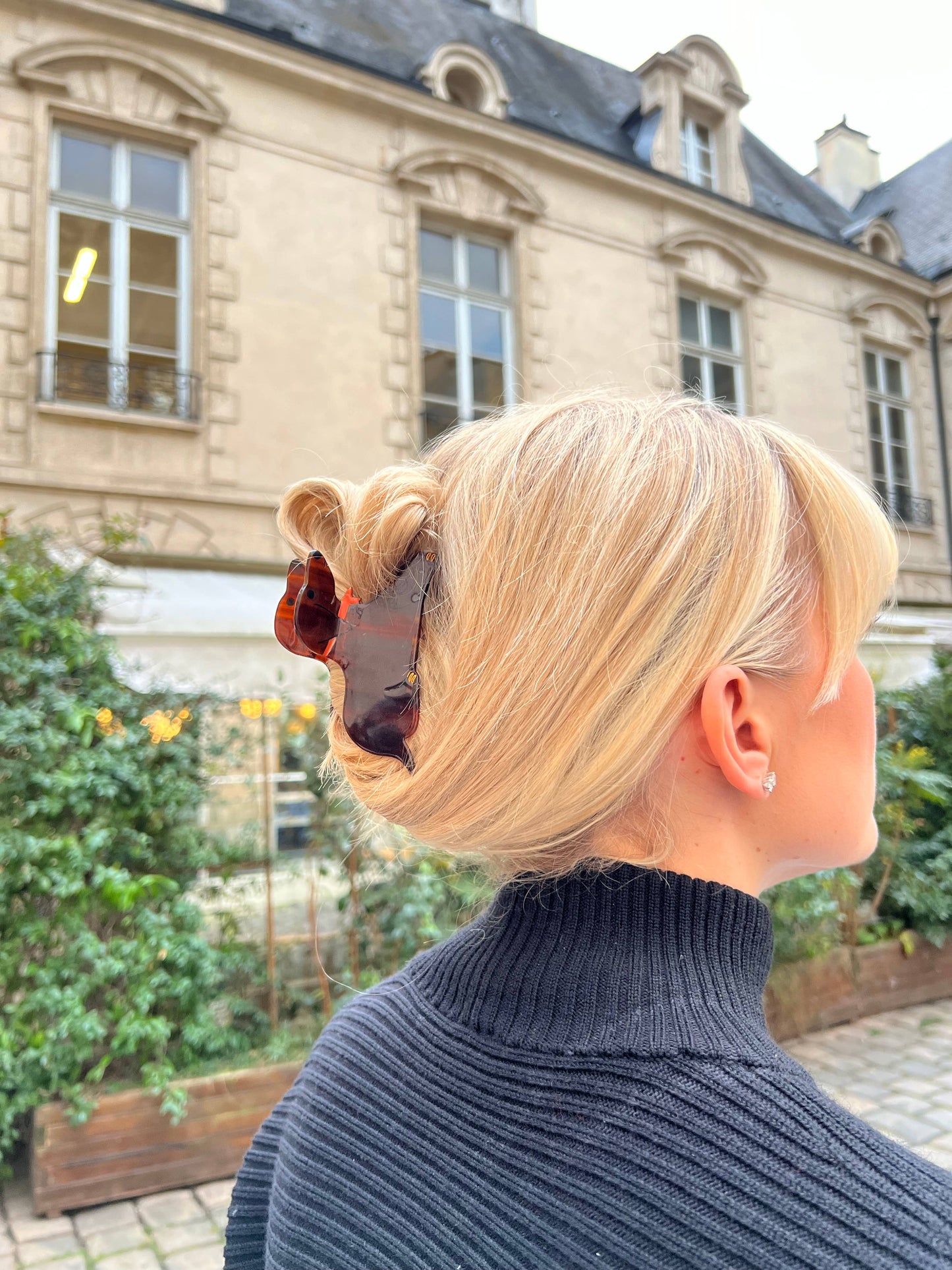 A woman with blonde hair styled in an elegant updo, secured with a dachshund-shaped hair claw. The tortoiseshell clip, featuring the playful silhouette of a dachshund with an orange bow, stands out against her hair. She is wearing a black ribbed turtleneck sweater, and the background features a classic building with large windows and detailed architecture.