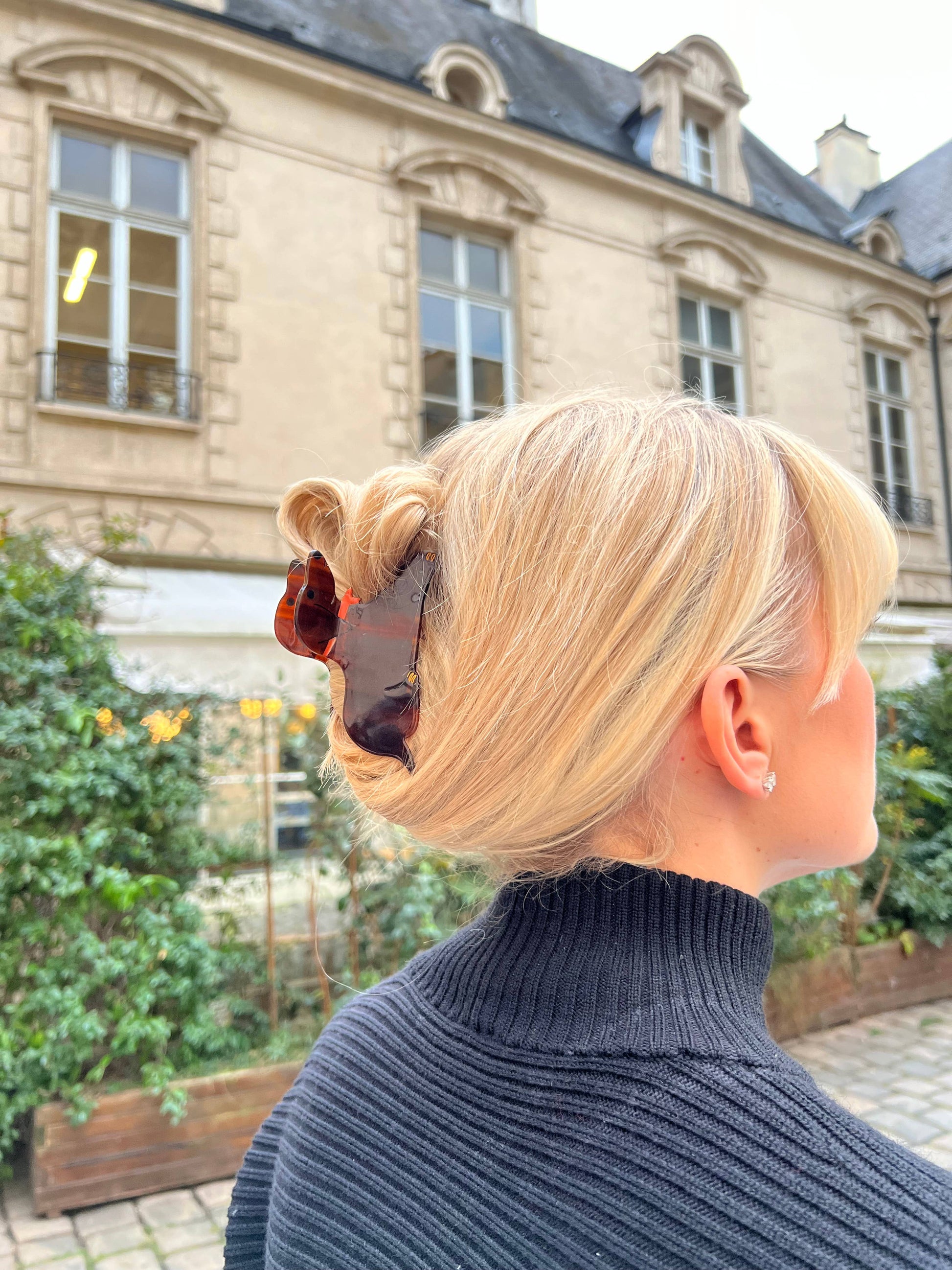 A woman with blonde hair styled in an elegant updo, secured with a dachshund-shaped hair claw. The tortoiseshell clip, featuring the playful silhouette of a dachshund with an orange bow, stands out against her hair. She is wearing a black ribbed turtleneck sweater, and the background features a classic building with large windows and detailed architecture.