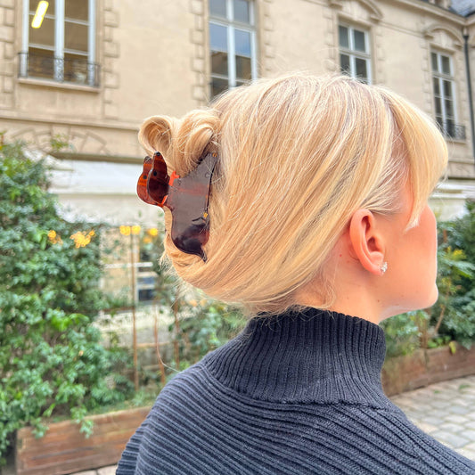 A woman with blonde hair styled in an elegant updo, secured with a dachshund-shaped hair claw. The tortoiseshell clip, featuring the playful silhouette of a dachshund with an orange bow, stands out against her hair. She is wearing a black ribbed turtleneck sweater, and the background features a classic building with large windows and detailed architecture.