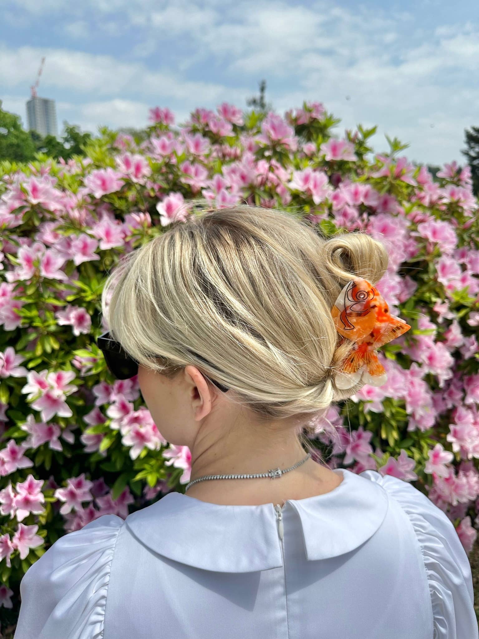A woman with blonde hair styled in a loose bun wears the goldfish-shaped hair claw, securing her hair with a pop of vibrant orange. She is dressed in a white puff-sleeve blouse and wears a delicate silver necklace. The background is filled with blooming pink flowers, creating a soft, romantic setting.