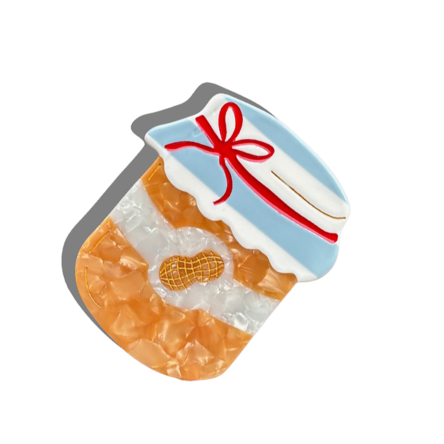 A peanut butter jar-shaped hair claw, featuring a marbled golden-brown body, a white label with a gold-etched peanut icon, and a blue and white gingham-style lid tied with a red bow. The clip is designed with a curved grip and gold detailing, casting a soft shadow against a white background.