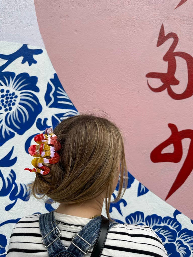  A person wearing the hand-painted snake-shaped hair claw clip in red, pink, and yellow. Their hair is styled in a loose updo, and they are standing against a vibrant mural featuring blue floral designs, a pink background, and red Chinese characters.