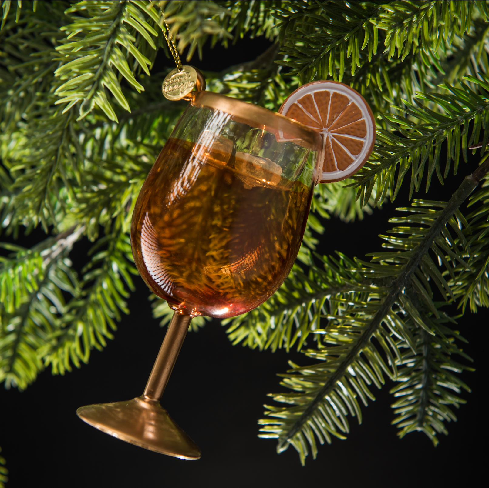 Hand-blown glass Aperol Spritz ornament featuring frosty ice and a hand-painted orange wheel garnish, hanging from a Christmas` tree branch