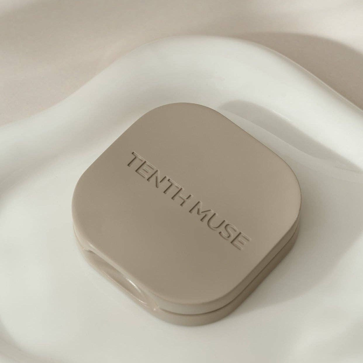 A clay square refillable compact for the tenth muse solid perfume balms. The compact is sitting on top of a white tray and has 'TENTH MUSE' lightly carved into the compact. 