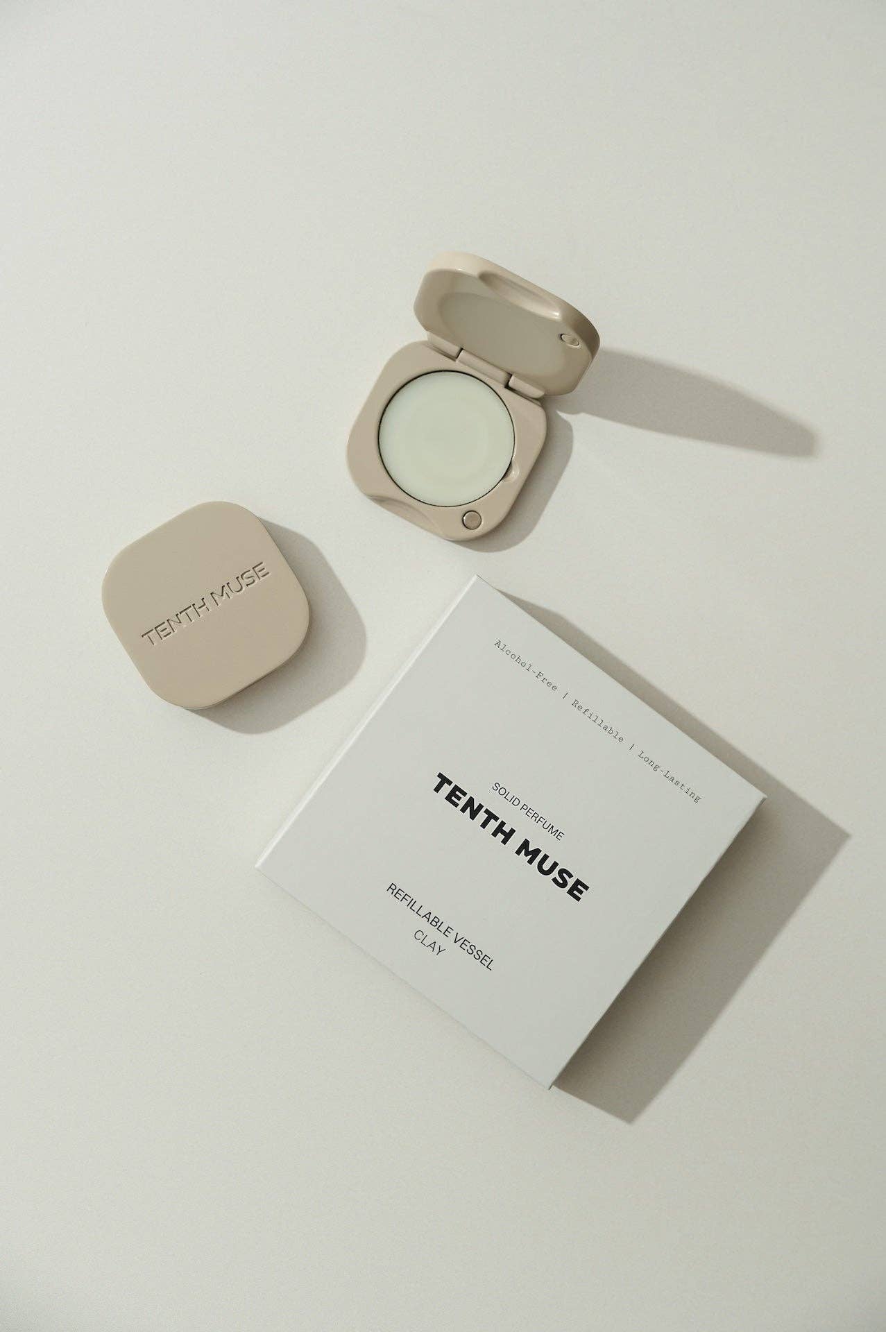 A clay square refillable compact for the tenth muse solid perfume balms that has 'TENTH MUSE' lightly carved into the compact sits next to an open compact that has a balm inside of it, beside a white square packaging box for the Tenth Muse refillable vessel. At the top of the box it says Alcohol-free, Refillable, Long-lasting'. Beneath that it says Solid Perfume and underneath in black box letters 'TENTH MUSE'. At the bottom of the box it says Refillable Vessel and Clay beneath, that being the shade. 