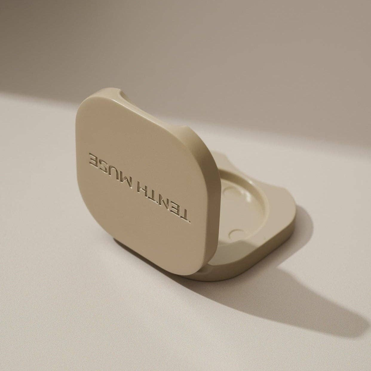 A halfway open clay square refillable compact for the tenth muse solid perfume balms that has 'TENTH MUSE' lightly carved into the compact and shows the hole in which the solid perfume balm goes inside of. 