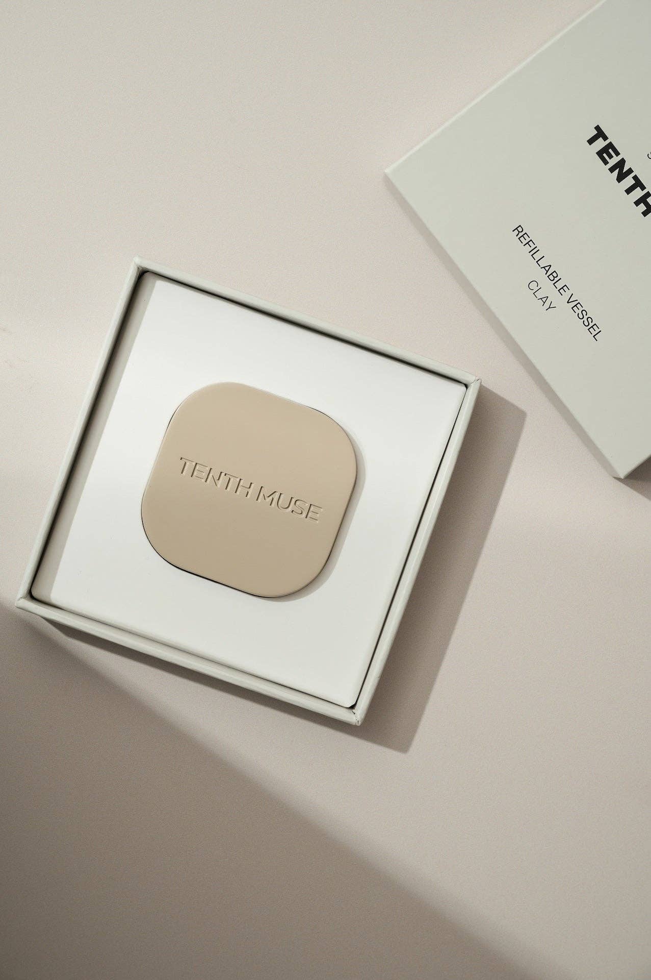 A clay square refillable compact for the tenth muse solid perfume balms. The compact is sitting inside of the packaging that the compact comes in which is a square box that is white on the inside and has 'TENTH MUSE' lightly carved into the compact. Next to the open box is the lid of the compact box which is also clay and says TENTH MUSE in black followed by Refillable Vessle and underneath that Clay, the shade of the compact. 
