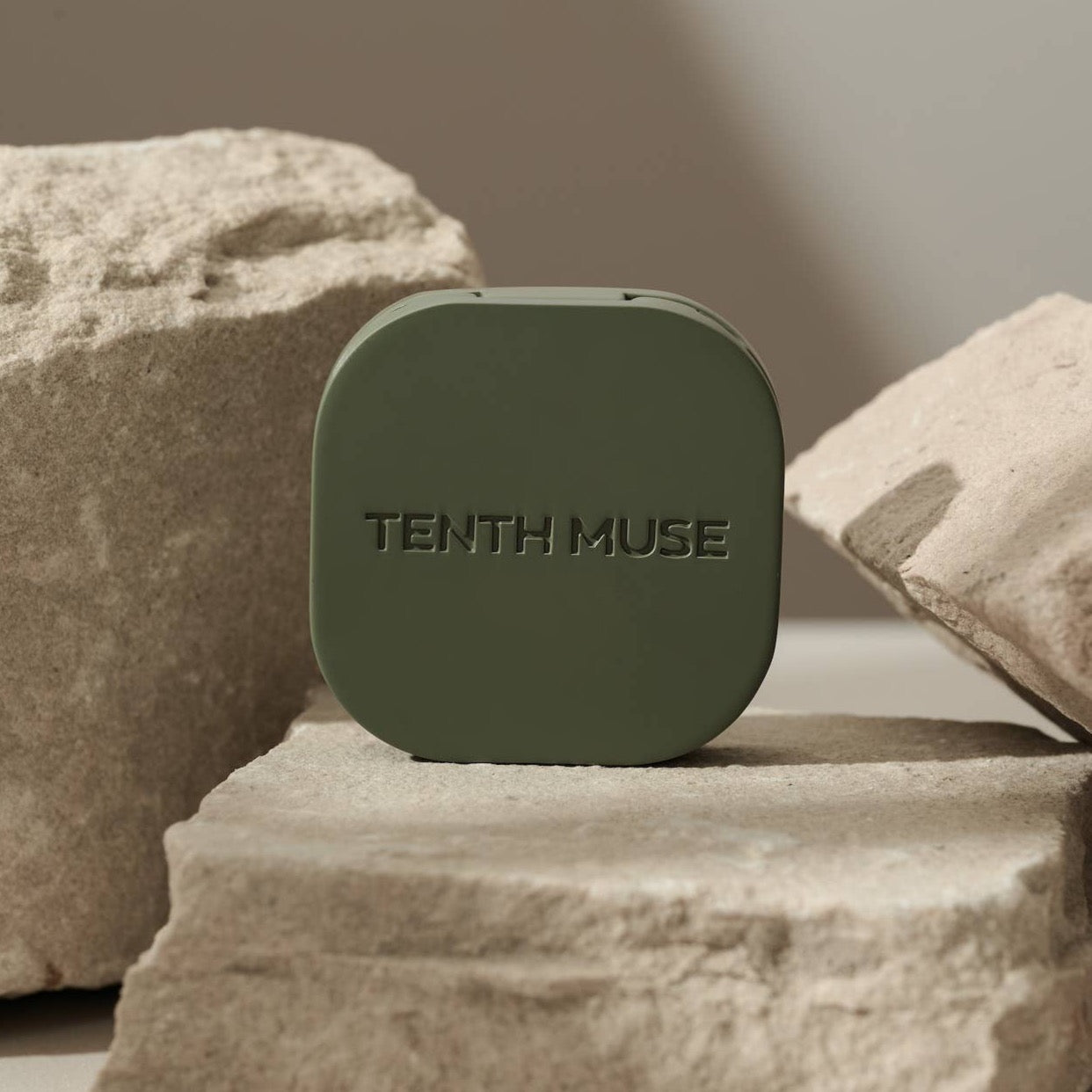 A green/matcha square refillable compact for the tenth muse solid perfume balms that has 'TENTH MUSE' lightly carved into the compact sits on top of a clay rock, with two on either side of the compact. 